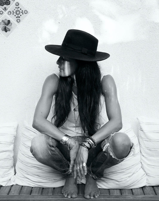 Mara Scalise seated cross-legged in a black hat, tank top, jeans, and layered jewelry