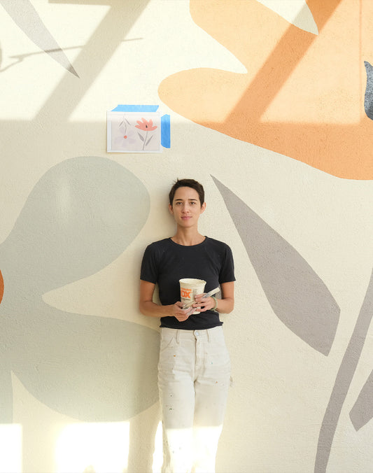 Liesel Plambeck holding a takeaway cup in front of a colorful abstract mural