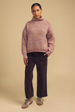 A person stands against a beige background wearing the DENNIE SWEATER from Velvet by Graham & Spencer, paired with brown wide-leg pants and beige sneakers, looking directly at the camera with a neutral expression. The cozy texture of the DENNIE SWEATER adds warmth and style to the ensemble.