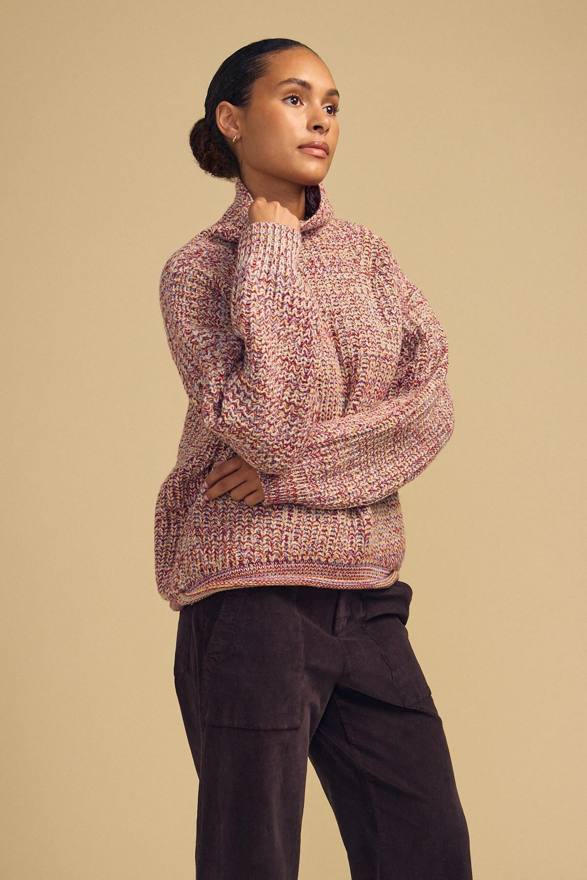 A person stands against a beige background wearing the DENNIE SWEATER by Velvet by Graham & Spencer—a marled chunky knit turtleneck—paired with dark pants, with one arm folded and the other hand resting near their chin.-42181521637569