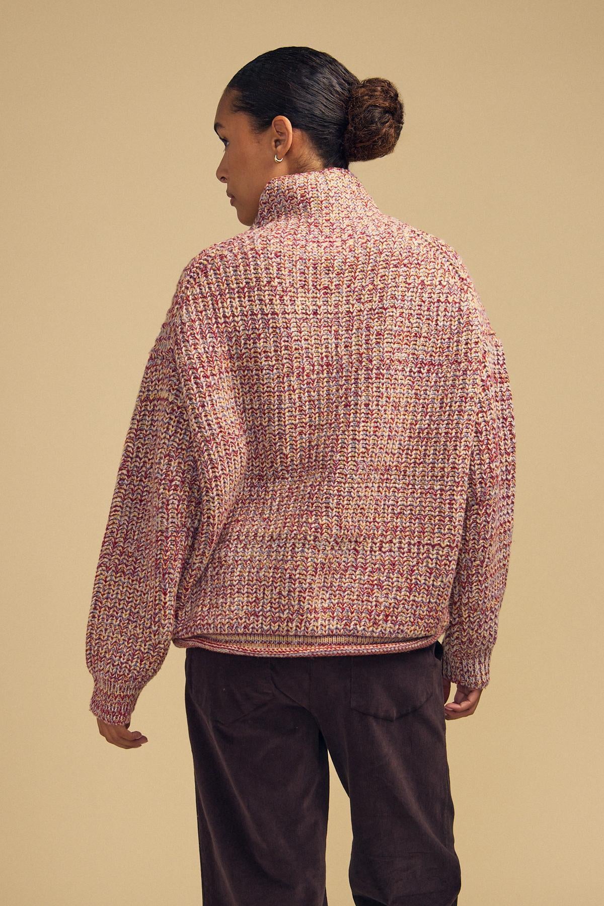 A person stands with their back to the camera, wearing the Velvet by Graham & Spencer DENNIE SWEATER—a chunky knit featuring a bulky, textured, multicolored design—paired with dark pants against a plain beige background.-42181521604801