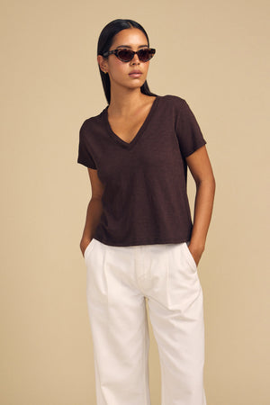 A model wearing a dark brown ELISHA TEE by Velvet by Graham & Spencer with white trousers and heart-shaped sunglasses.