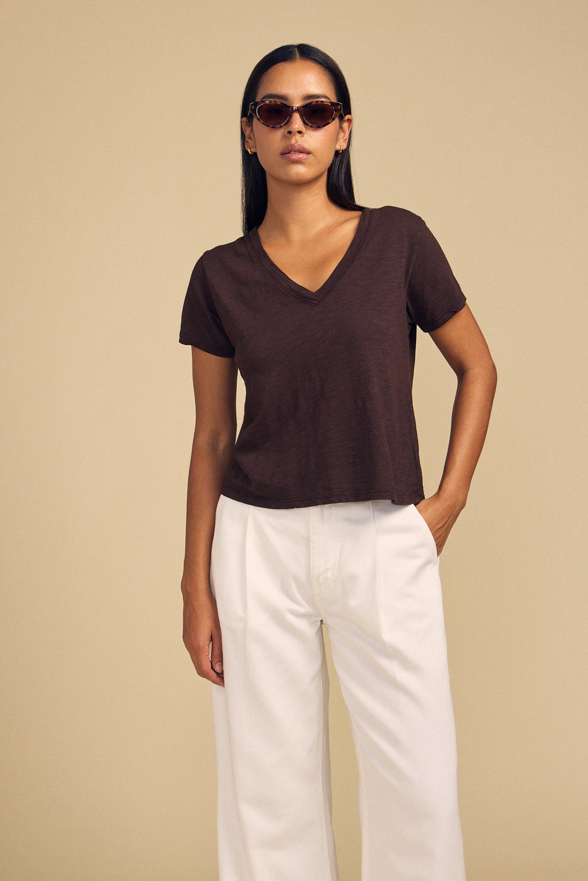 A woman wearing a dark brown cotton slub V-neck ELISHA TEE by Velvet by Graham & Spencer and white trousers, posing with her hand in her pocket.
