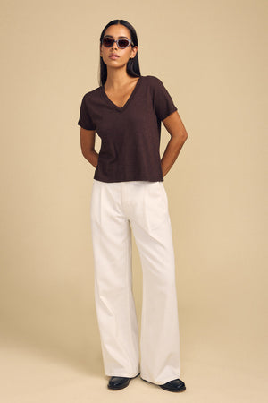 A person wearing a dark brown cotton slub V-neck ELISHA TEE by Velvet by Graham & Spencer and white wide-leg trousers.