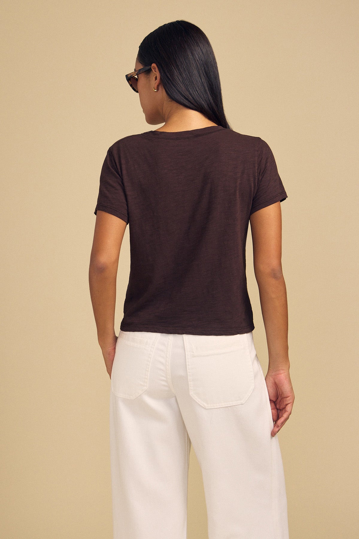 Back view of a person wearing a dark brown ELISHA TEE in cotton slub with a crew neck and white pants, a timeless wardrobe staple.-46505564700865