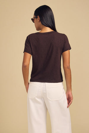 Back view of a person wearing a dark brown ELISHA TEE in cotton slub with a crew neck and white pants, a timeless wardrobe staple.