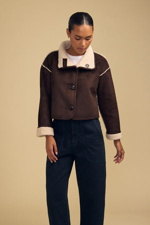 A person wears the ESME FAUX SHERPA REVERSIBLE JACKET by Velvet by Graham & Spencer—a brown, cropped design—layered over a white shirt and paired with dark high-waisted pants, standing against a beige background.