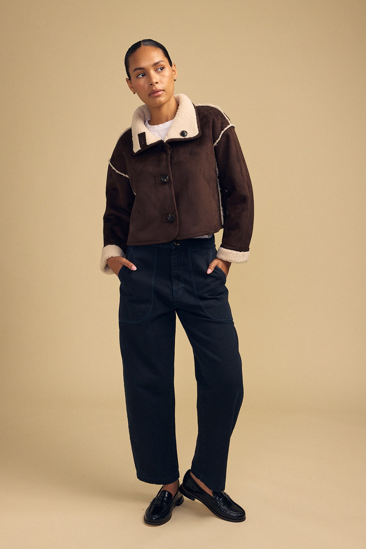 A person stands against a beige background wearing the ESME FAUX SHERPA REVERSIBLE JACKET by Velvet by Graham & Spencer, paired with dark loose-fitting trousers and black shoes, with hands in pockets and a neutral facial expression.