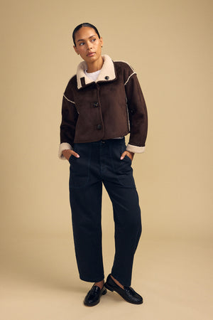 A person stands against a beige background wearing the ESME FAUX SHERPA REVERSIBLE JACKET by Velvet by Graham & Spencer, paired with dark loose-fitting trousers and black shoes, with hands in pockets and a neutral facial expression.