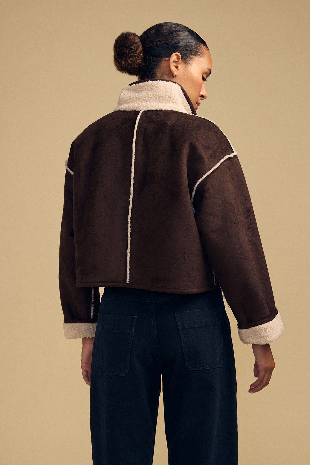 A person faces away from the camera, wearing the ESME FAUX SHERPA REVERSIBLE JACKET by Velvet by Graham & Spencer—a brown faux suede sherpa jacket with white trim—paired with dark pants, set against a plain beige background.
