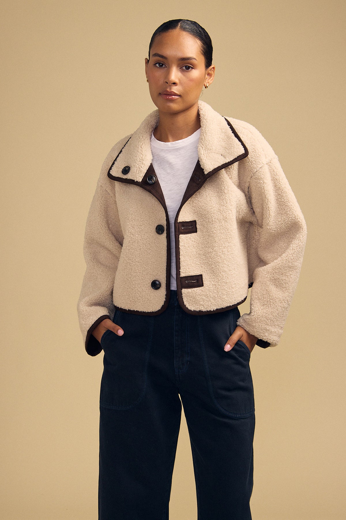 A person wearing the ESME FAUX SHERPA REVERSIBLE JACKET by Velvet by Graham & Spencer, featuring a cream-colored faux sherpa texture and large buttons, layered over a white shirt and dark pants, stands against a plain tan background.