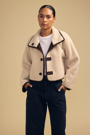 A person wearing the ESME FAUX SHERPA REVERSIBLE JACKET by Velvet by Graham & Spencer, featuring a cream-colored faux sherpa texture and large buttons, layered over a white shirt and dark pants, stands against a plain tan background.
