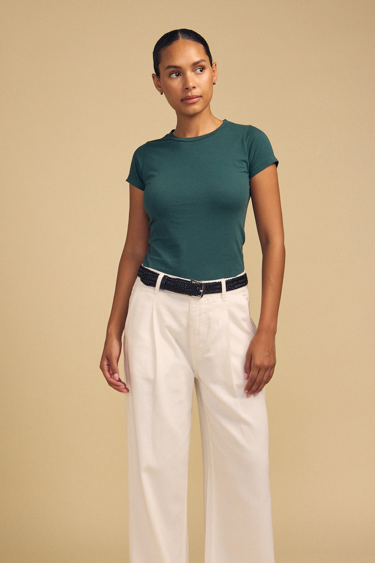 A person stands against a beige background wearing the Velvet by Graham & Spencer JEMMA TEE in ultra-soft dark green, paired with cream wide-leg pants and a black belt.