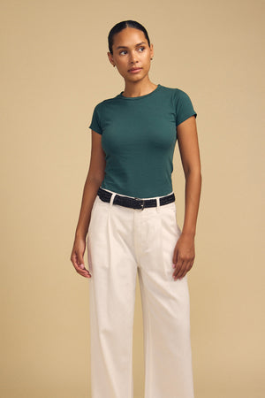 A person stands against a beige background wearing the Velvet by Graham & Spencer JEMMA TEE in ultra-soft dark green, paired with cream wide-leg pants and a black belt.