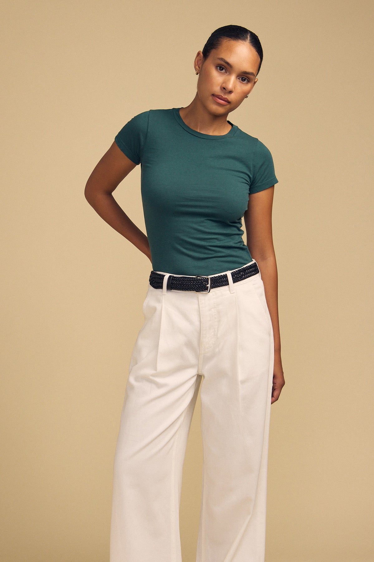 A person stands against a beige background wearing the Velvet by Graham & Spencer JEMMA TEE in ultra-soft green, paired with white high-waisted pants and a black belt, with one hand behind their back.-42182013485249