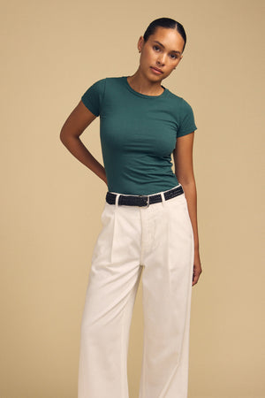 A person stands against a beige background wearing the Velvet by Graham & Spencer JEMMA TEE in ultra-soft green, paired with white high-waisted pants and a black belt, with one hand behind their back.