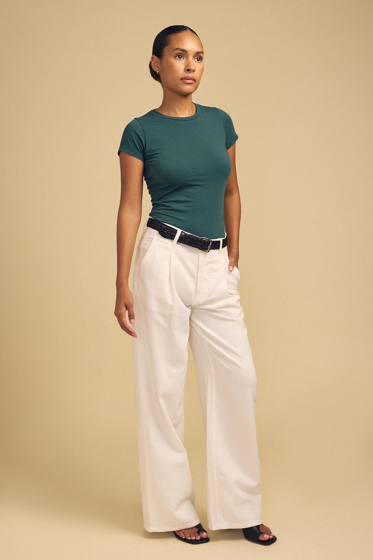 A person stands against a beige background wearing the ultra-soft JEMMA TEE in green from Velvet by Graham & Spencer, styled with wide-leg white trousers, a black belt, and black sandals.-42182013452481
