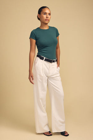 A person stands against a beige background wearing the ultra-soft JEMMA TEE in green from Velvet by Graham & Spencer, styled with wide-leg white trousers, a black belt, and black sandals.