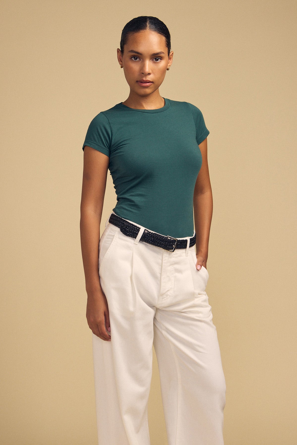 Standing against a beige background, the person wears the JEMMA TEE by Velvet by Graham & Spencer—an ultra-soft fitted green crew neck—paired with white high-waisted trousers and a black belt. Their hair is pulled back, and their expression is neutral.