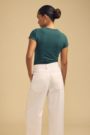 A person stands facing away against a beige background, wearing the ultra-soft JEMMA TEE by Velvet by Graham & Spencer in green, paired with high-waisted white pants.
