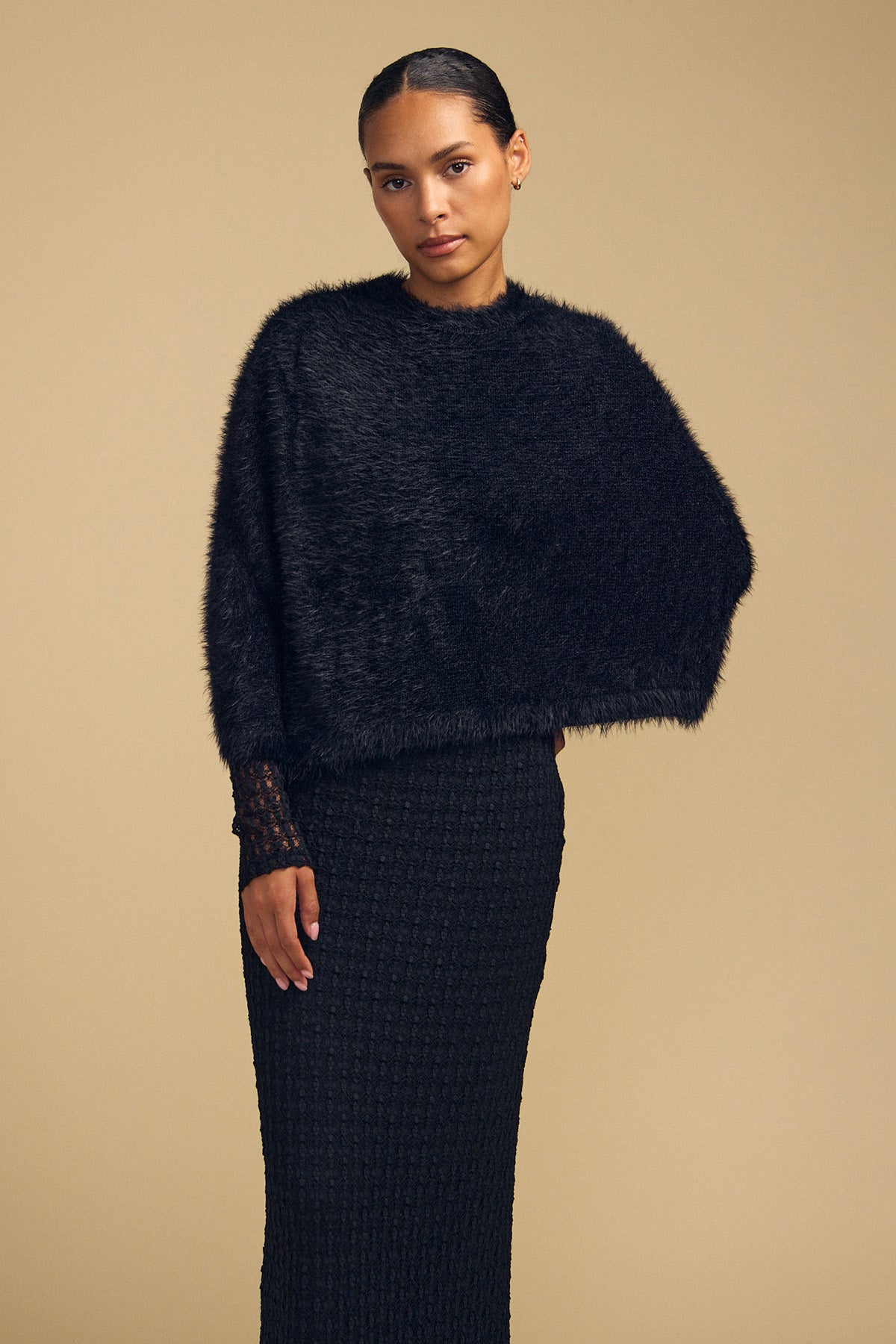 A model wears a black, fuzzy LAYLA FEATHER YARN CAPE SWEATER over a matching textured skirt.-46488054890689
