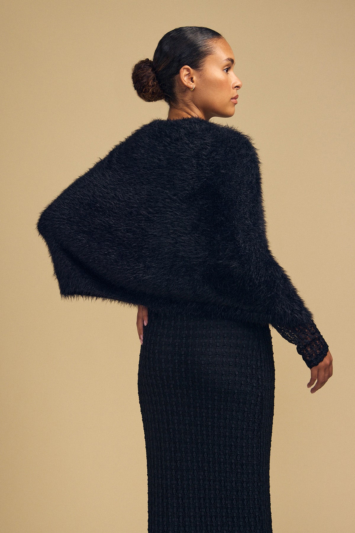 A person wearing a fuzzy black LAYLA FEATHER YARN CAPE SWEATER over a black textured dress.-46488054792385