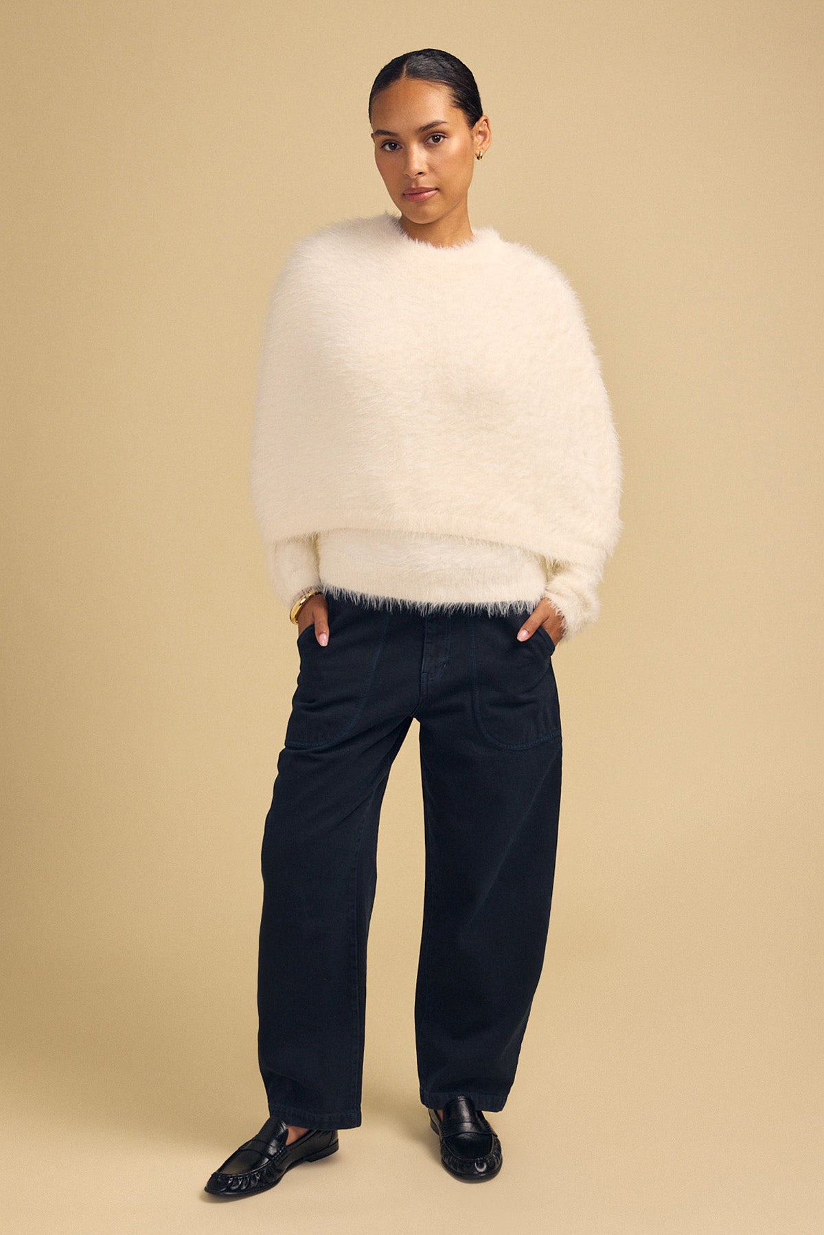 A person wearing a cream-colored, fuzzy LAYLA FEATHER YARN CAPE SWEATER with dark trousers and black loafers.-46488054759617