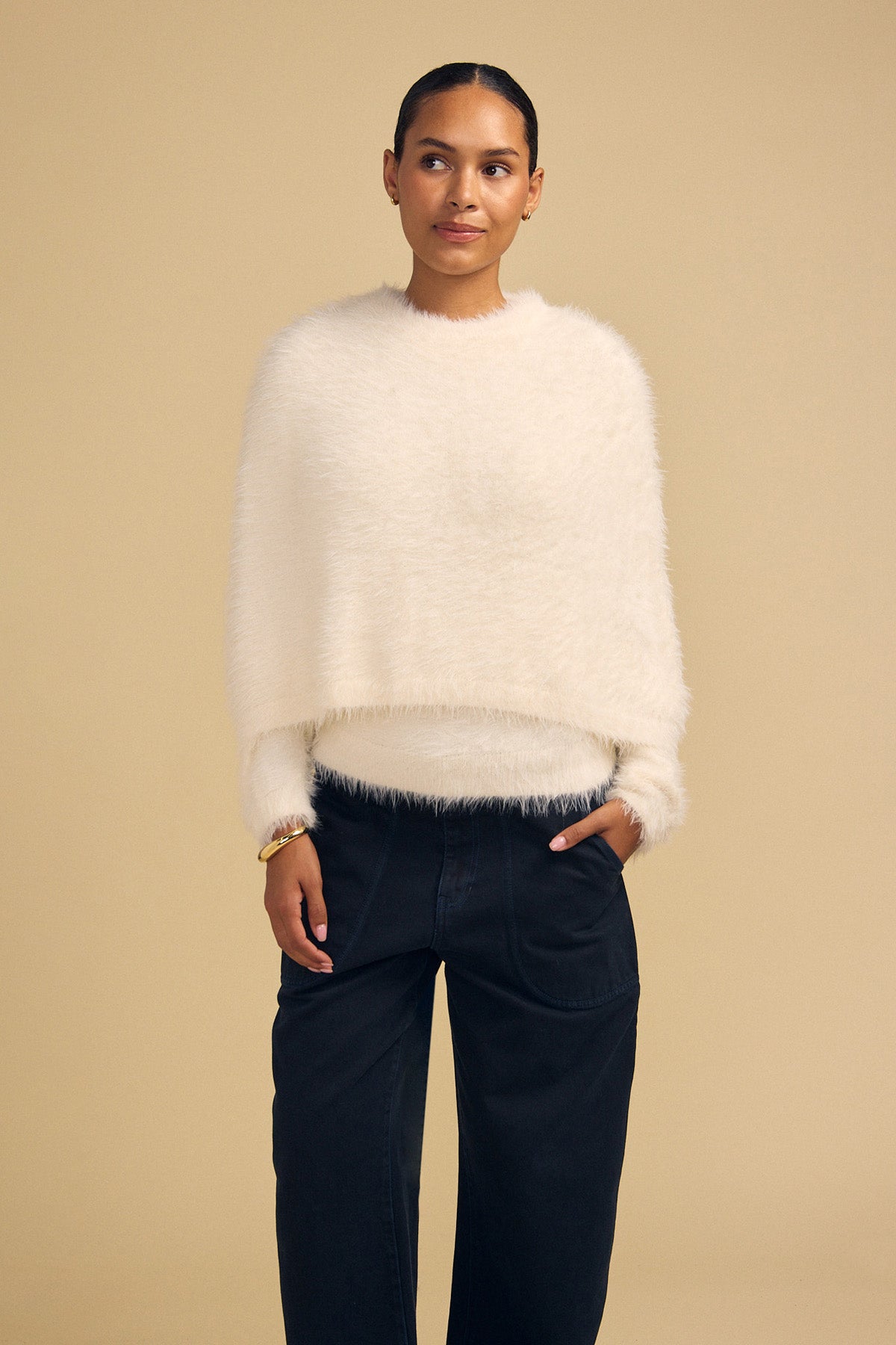 A person wearing a cream-colored, fuzzy, cropped LAYLA FEATHER YARN CAPE SWEATER with long sleeves and dark wide-leg trousers.