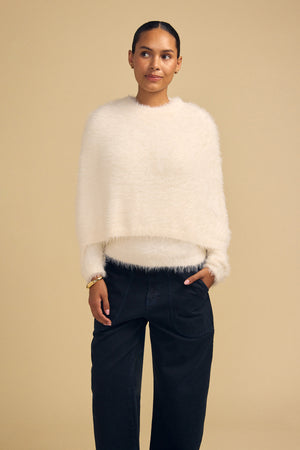 A person wearing a cream-colored, fuzzy, cropped LAYLA FEATHER YARN CAPE SWEATER with long sleeves and dark wide-leg trousers.