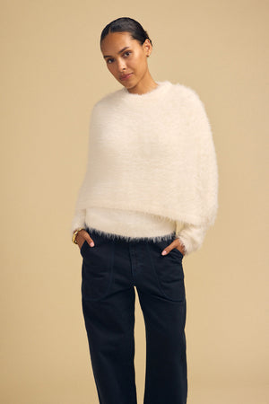 A model wearing a cream-colored, fuzzy LAYLA FEATHER YARN CAPE SWEATER and dark trousers.