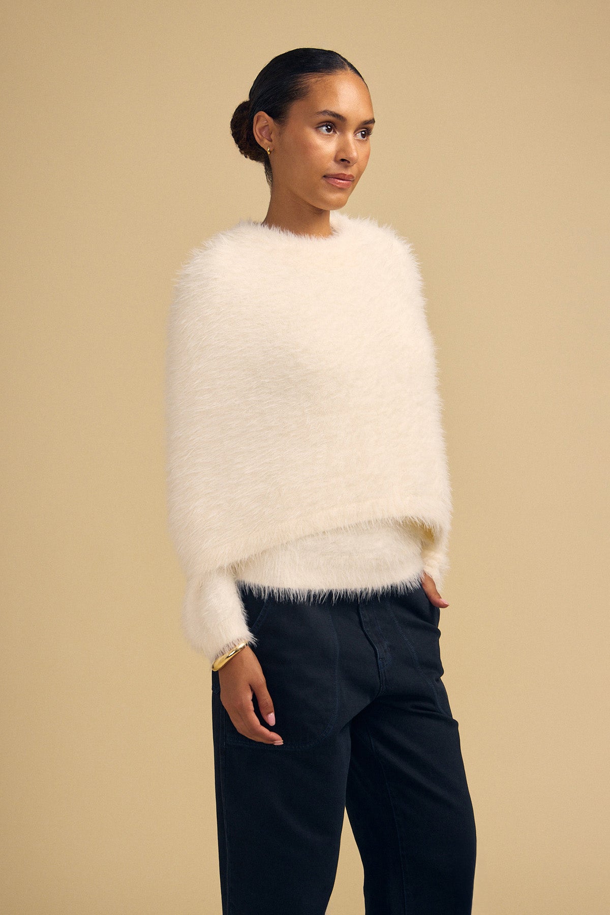 A person wears a cream-colored, fuzzy, cropped LAYLA FEATHER YARN CAPE SWEATER with long sleeves over dark trousers.-46488054563009