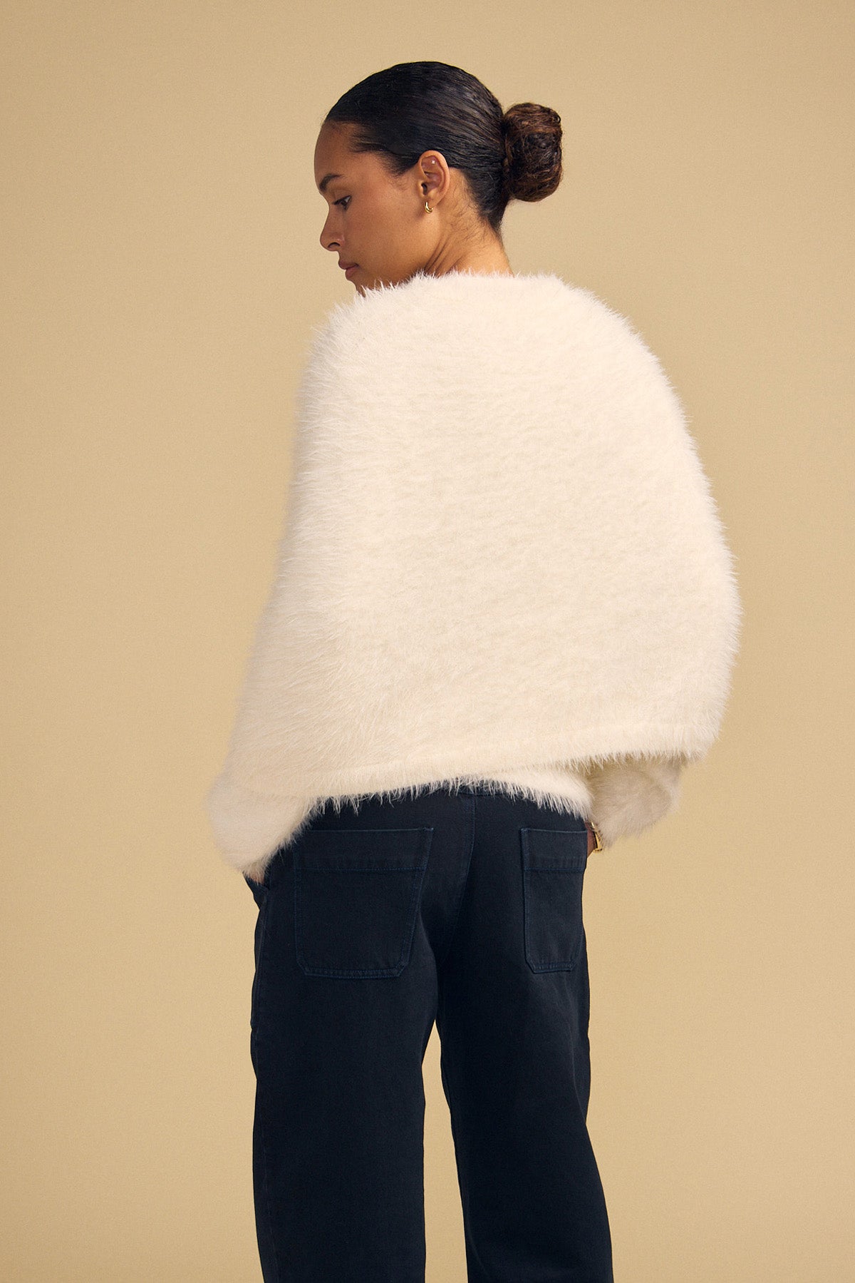 A person wearing a fuzzy, cream-colored LAYLA FEATHER YARN CAPE SWEATER and dark trousers.