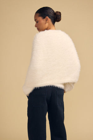 A person wearing a fuzzy, cream-colored LAYLA FEATHER YARN CAPE SWEATER and dark trousers.