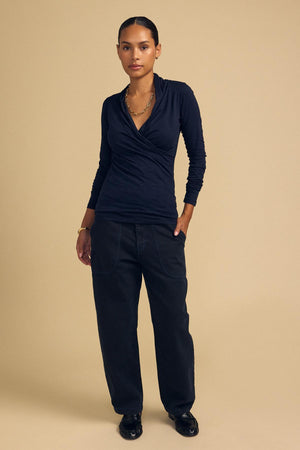 A person stands against a beige background wearing the MERI TEE by Velvet by Graham & Spencer, styled with loose black pants, black shoes, and gold jewelry.
