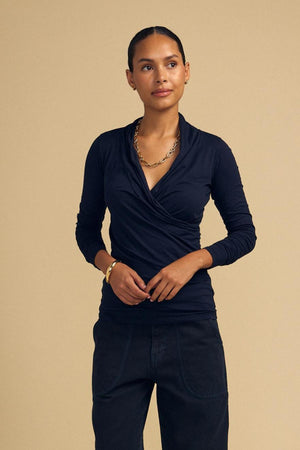 Wearing the MERI TEE by Velvet by Graham & Spencer, a person in a black V-neck wrap tee, gold chain necklace, and dark pants poses against a beige background.