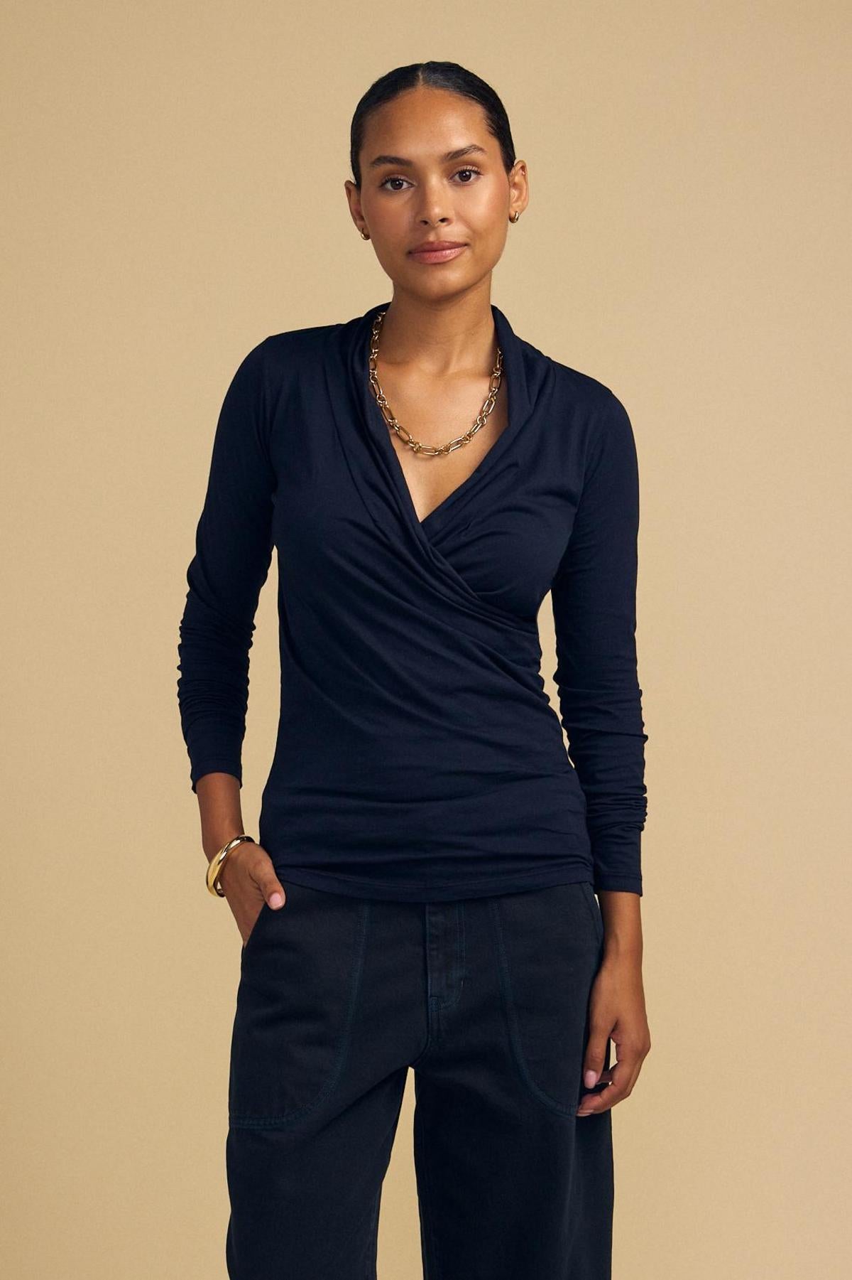 A person stands against a beige background wearing the MERI TEE by Velvet by Graham & Spencer, black pants, a gold chain necklace, and a gold bracelet.