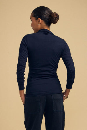 Person with dark hair in a bun wears the MERI TEE by Velvet by Graham & Spencer and black pants, standing with their back to the camera against a plain tan background.