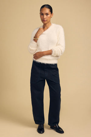 A person wears a fuzzy, cream-colored PENNY FEATHER YARN SWEATER with a V-neck, paired with dark blue wide-leg jeans and black loafers for versatile comfort.