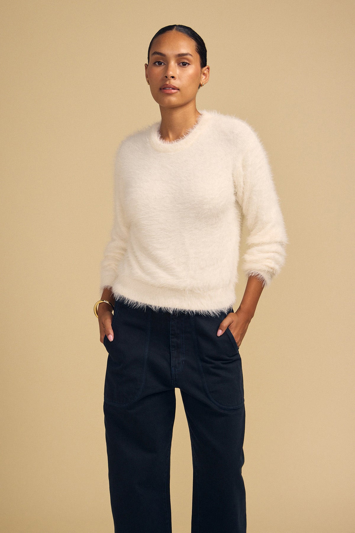 A person wears a cream-colored, fuzzy long-sleeved PENNY FEATHER YARN SWEATER by Velvet by Graham & Spencer, featuring a crew neckline and dark pants.-46488052433089