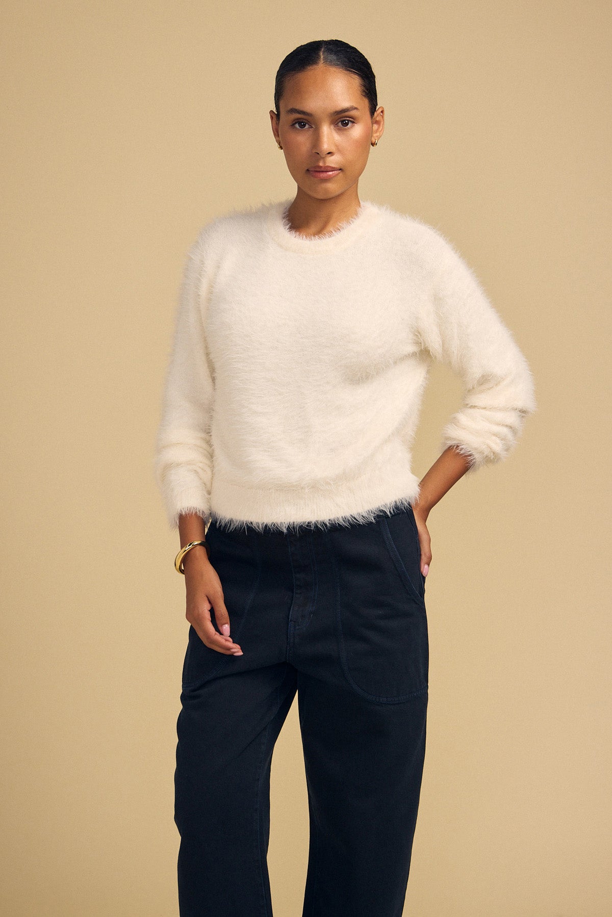 A woman wearing a cream-colored, fuzzy long-sleeved PENNY FEATHER YARN SWEATER by Velvet by Graham & Spencer with a classic crew neckline and dark trousers.