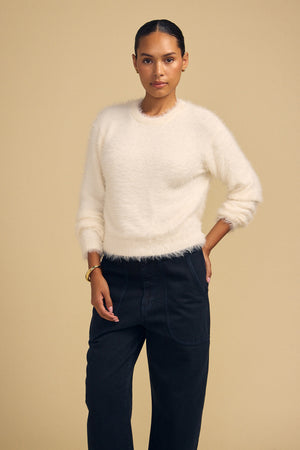 A woman wearing a cream-colored, fuzzy long-sleeved PENNY FEATHER YARN SWEATER by Velvet by Graham & Spencer with a classic crew neckline and dark trousers.