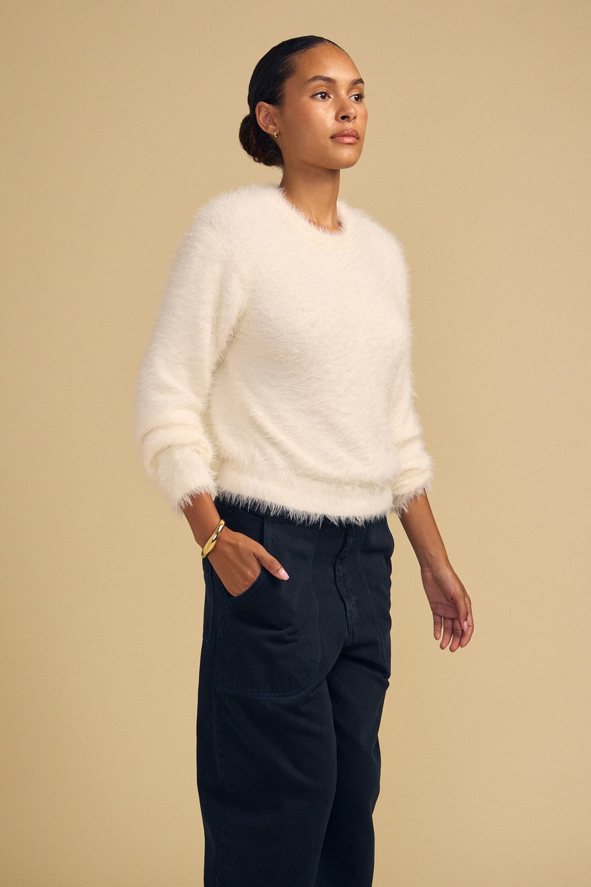 A model wears a cream-colored, fuzzy, long-sleeved PENNY FEATHER YARN SWEATER by Velvet by Graham & Spencer with a classic crew neckline and dark pants, with her hand in her pocket.