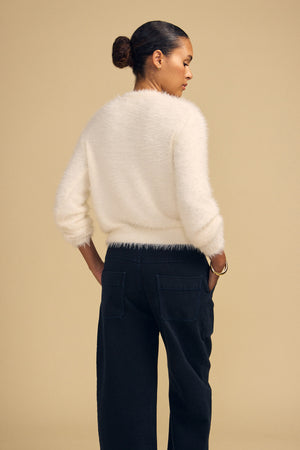 A person seen from behind wears a white fuzzy long-sleeved PENNY FEATHER YARN SWEATER by Velvet by Graham & Spencer with a classic crew neckline and dark pants, hands tucked into their pockets.