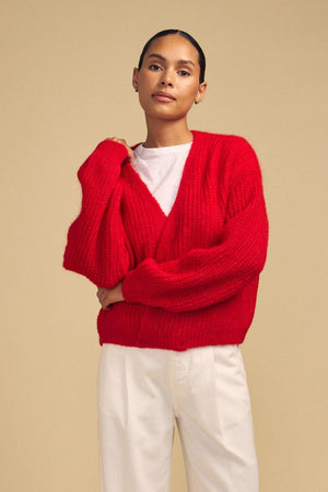 A person stands against a beige background, wearing a white shirt, white pants, and the RANYA ALPACA CARDIGAN by Velvet by Graham & Spencer.