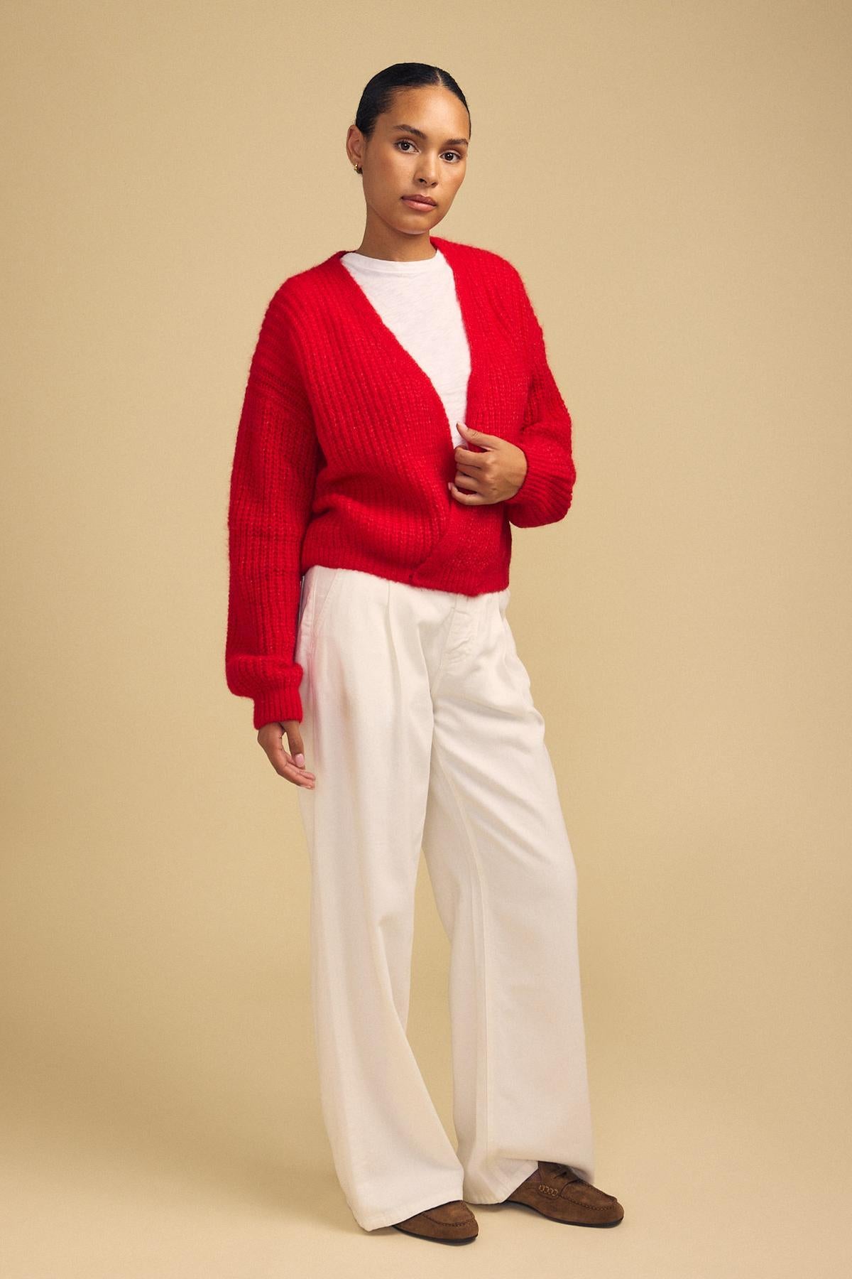 A person stands against a beige background, wearing the RANYA ALPACA CARDIGAN in red by Velvet by Graham & Spencer over a white shirt, paired with white wide-leg pants and brown shoes.-42181908988097
