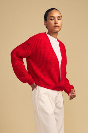 A person stands against a plain beige background, wearing the RANYA ALPACA CARDIGAN by Velvet by Graham & Spencer—a cozy red layering sweater styled over a white shirt and cream pants.