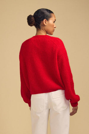 A person stands with their back to the camera, wearing the Velvet by Graham & Spencer RANYA ALPACA CARDIGAN in a rich red textured knit paired with white pants, set against a beige background.