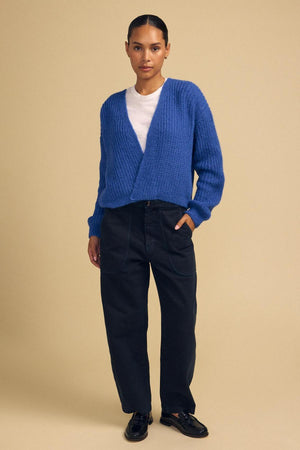 Standing against a beige background, the person is wearing the RANYA ALPACA CARDIGAN by Velvet by Graham & Spencer in blue, layered over a white shirt and paired with black high-waisted pants and black shoes for a cozy look.