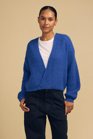 A person stands against a plain beige background, wearing the Velvet by Graham & Spencer RANYA ALPACA CARDIGAN—a blue alpaca blend piece—layered over a white T-shirt and paired with dark pants for a cozy look.