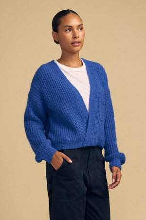 A person wearing the RANYA ALPACA CARDIGAN by Velvet by Graham & Spencer—a bright blue alpaca blend piece—over a white shirt and black pants stands against a plain beige background.